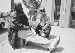 Street person siting next to a pole selling a newspaper put out by homeless people, while worker Terry Kalahar sits by him.